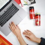 Top view of a workspace with hands on a laptop and Coca-Cola cans nearby.