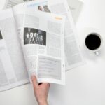 A close-up shot of a hand holding a newspaper with a coffee cup on a white surface.
