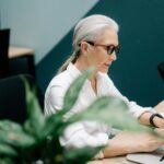 Confident senior woman with glasses working on a laptop in a modern office setting.