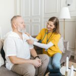 A nurse uses a stethoscope for a home check-up on a senior adult in a cozy living room.