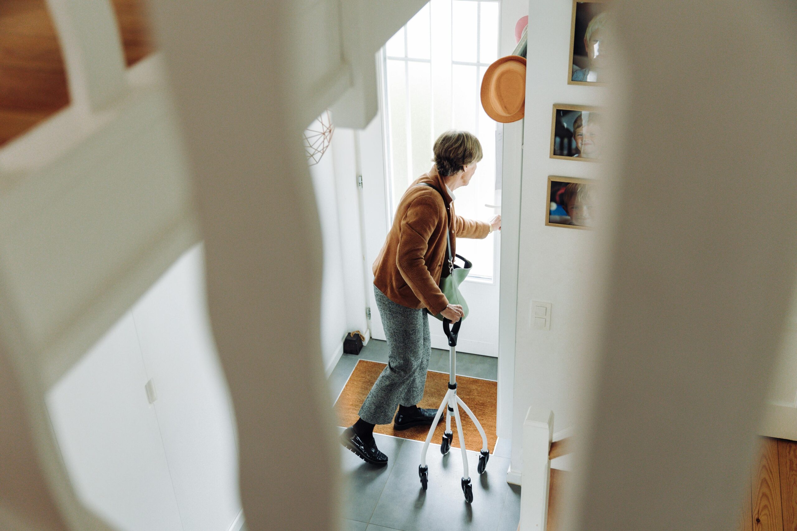Senior woman using rollator indoors near doorway, preparing to leave home.